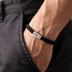 Puede incluir: Pulsera trenzada negra con una barra plateada con las letras "S", un símbolo de corazón y la letra "W". La pulsera se lleva en la muñeca, con el brazo de la persona y un pantalón gris oscuro en el fondo.