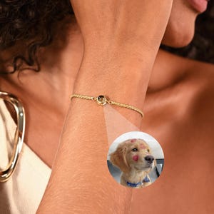May include: A delicate gold bracelet with a small, round charm. The bracelet is on a person's wrist. A circular inset shows a golden retriever puppy with red heart-shaped kisses on its face.