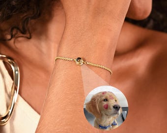 以下が含まれることがあります： 小さな丸いチャームが付いた繊細なゴールドのブレスレット。ブレスレットは人の手首にあります。円形のインセットには、顔に赤いハート型のキスをしたゴールデンレトリバーの子犬が表示されています。