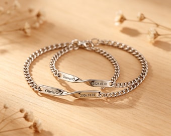 May include: Two silver-colored chain bracelets. Each bracelet has a rectangular plate with the names "Olivia" and "Thomas" engraved, along with a heart symbol. The date "2026.01.01" is also engraved on each plate.