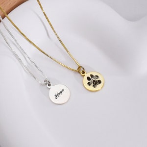 May include: Two delicate necklaces. One is silver with the name "Hugo" engraved on a round pendant. The other is gold, featuring a paw print on a similar pendant. Both have fine chain links and are displayed on a white surface.