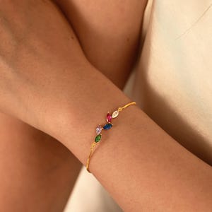 Puede incluir: Una pulsera dorada con una cadena delicada y una serie de gemas de colores con corte marquesa. Las gemas están dispuestas en un patrón lineal, con piedras rojas, blancas, rosas, azules y verdes. La pulsera se lleva en la muñeca.