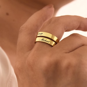 Può includere: Un anello color oro con i nomi "Amaya" e "Michael" incisi in caratteri neri. L'anello è indossato su un dito, mostrando la sua superficie lucida e il suo design personalizzato. L'anello è un semplice stile a fascia.