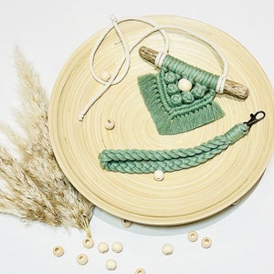 May include: A green macrame wall hanging with a wooden dowel and white beads. The hanging is on a wooden tray with a white cord and wooden beads.
