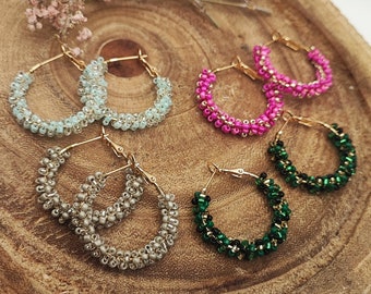 Pode incluir: Brincos de argola com contas em várias cores: azul claro, prateado, rosa e verde. Os brincos são exibidos numa tábua de madeira com flores secas.