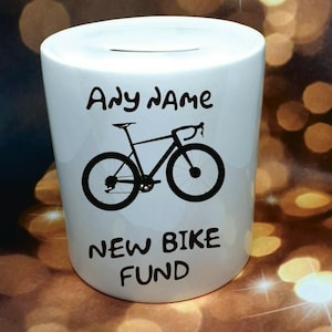 Może przedstawiać: Biała ceramiczna skarbonka z czarną sylwetką roweru i tekstem "Any name New Bike Fund".