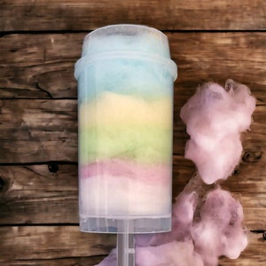 May include: A clear plastic container filled with layers of pastel-coloured cotton candy. The container has a handle and is sitting on a wooden surface. There are also loose pieces of cotton candy in pink and white.