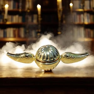 May include: A golden Snitch prop from the Harry Potter series, with detailed wing and sphere design. The prop is set on a wooden surface, with a background of candles and bookshelves, creating a magical atmosphere.