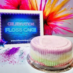 May include: A multi-coloured cotton candy cake with the words "Celebration Floss Cake" on a neon sign behind it. The cake is in a clear plastic container and is sitting on a white surface with purple and green sprinkles.