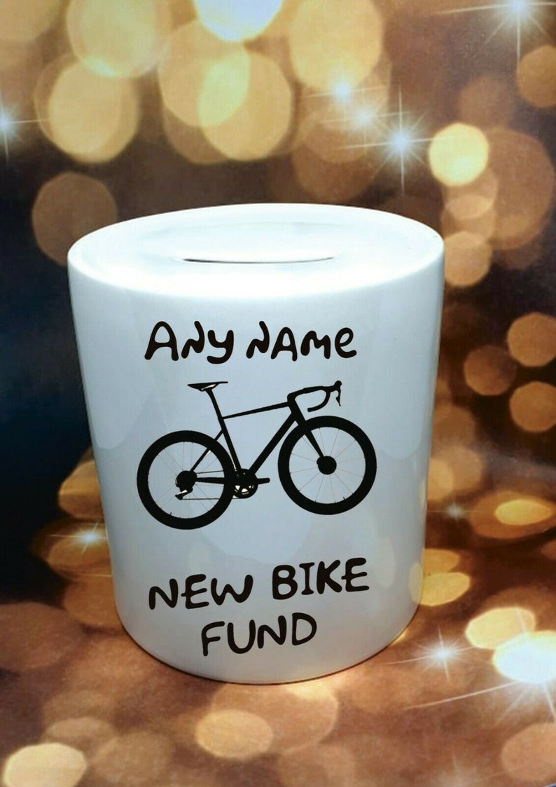 Może przedstawiać: Biała ceramiczna skarbonka z czarną sylwetką roweru i tekstem "Any name New Bike Fund".