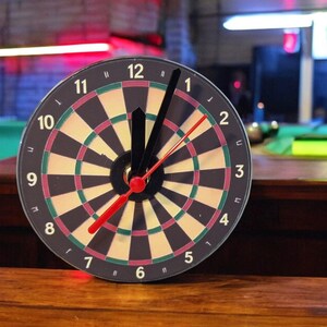 Op de afbeelding: Een dartbordklok met een zwart frame en een kleurrijk dartbordontwerp. De wijzers zijn zwart en rood en geven de tijd aan. De klok staat op een houten oppervlak, met een pooltafel op de achtergrond.