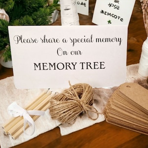 Könnte beinhalten: Ein Schild mit der Aufschrift "Bitte teile eine besondere Erinnerung an unserem GEDÄCHTNISBAUM". Auf einem Tisch liegen braune Papieretiketten, Schnur, Bleistifte und eine kleine Vase mit getrockneten Blumen.