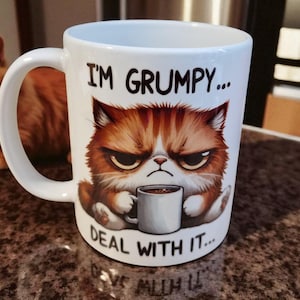 Op de afbeelding: Witte keramische mok met een illustratie van een chagrijnige kat die een kleine koffiekop vasthoudt. De mok heeft de tekst "I'M GRUMPY... DEAL WITH IT...". Op de achtergrond is een oranje tijgerkat te zien.