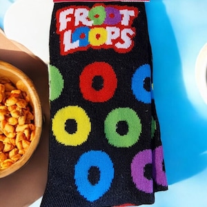 May include: Black crew socks with a pattern of colourful circles in red, yellow, green, blue, and purple. The socks display the text "Froot Loops" in white and red. The packaging states the socks fit men's shoe sizes 6-12.