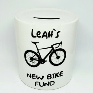 Może przedstawiać: Biała ceramiczna skarbonka z czarnym wzorem roweru i tekstem "Leah's New Bike Fund".