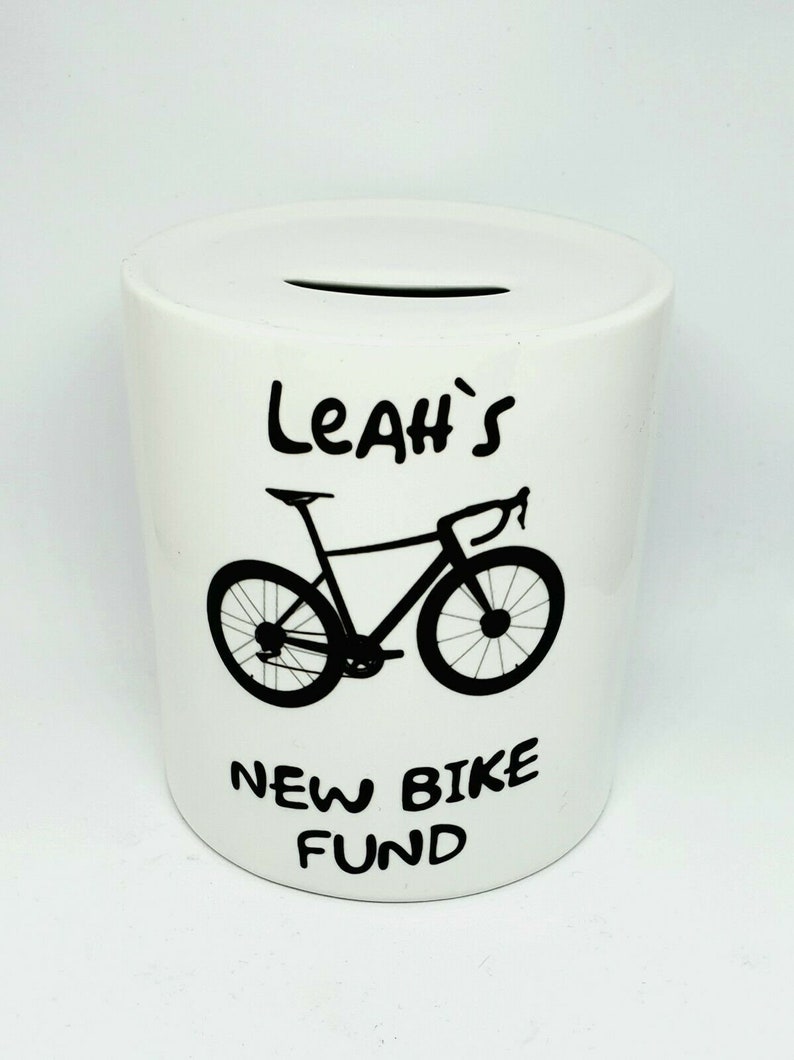 Może przedstawiać: Biała ceramiczna skarbonka z czarnym wzorem roweru i tekstem "Leah's New Bike Fund".