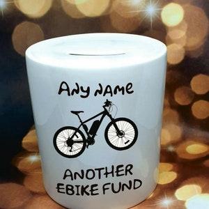 Może przedstawiać: Biała ceramiczna skarbonka z czarną sylwetką roweru elektrycznego i tekstem "Any name Another Ebike Fund".