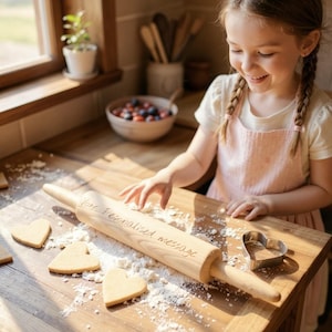 Può includere: Un mattarello di legno con la scritta "Your Personalised message" è su una superficie di legno con farina e biscotti a forma di cuore. È presente anche un tagliabiscotti a forma di cuore. Una persona giovane con un grembiule rosa sorride.