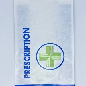 May include: A white paper bag featuring the blue text PRESCRIPTION printed vertically on the left side. A green cross icon inside a blue circle is centered on the bag. A blue horizontal band at the bottom contains the white text HANDLE MEDICINES WITH CARE.
