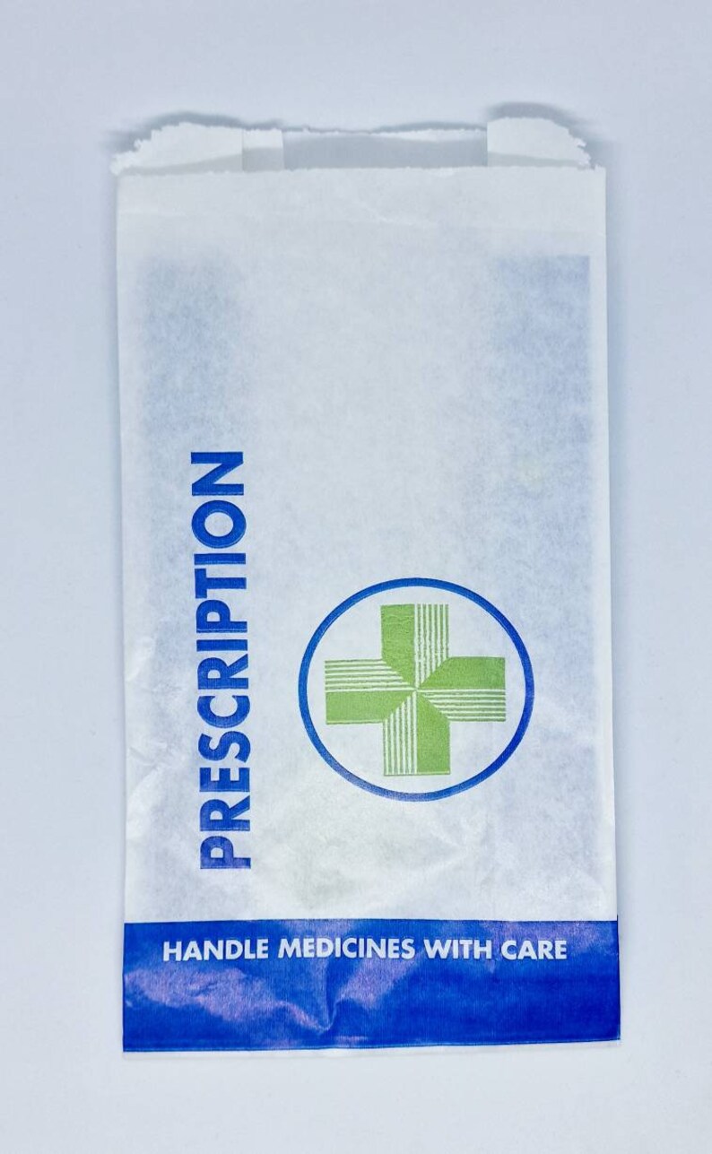 May include: A white paper bag featuring the blue text PRESCRIPTION printed vertically on the left side. A green cross icon inside a blue circle is centered on the bag. A blue horizontal band at the bottom contains the white text HANDLE MEDICINES WITH CARE.
