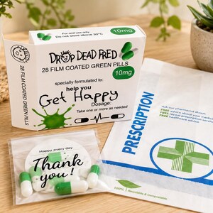 May include: A white rectangular box labeled Drop Dead Fred 28 film coated green pills 10mg with a green splash graphic, a small clear plastic bag containing green and white capsules with a Thank You tag, and a white prescription paper bag.