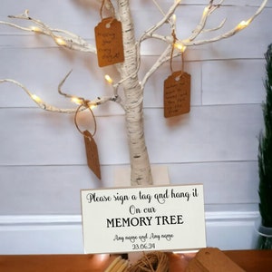 May include: A white birch tree with lights and brown paper tags hanging from the branches. A sign at the base reads "Please sign a tag and hang it on our MEMORY TREE Any name and Any name 23.06.24". There are also pencils, twine, and a stack of brown paper tags.