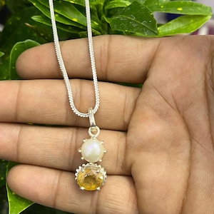 May include: A silver chain necklace with a pendant featuring a yellow gemstone and a white pearl. The pendant is set in a silver frame with a decorative design.