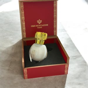 Op de afbeelding: Een witte parfumfles met een gouden dop en de tekst "Pomme d'Amour" op een label, gepresenteerd in een rode doos met gouden randen. De doos heeft de tekst "Gris Montaigne Paris" aan de binnenkant.