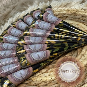Könnte beinhalten: Eine Sammlung von Henna-Kegeln, jeweils in schwarzes und goldfarbenes Papier mit einem Blattmuster gewickelt. Jeder Kegel hat ein Etikett mit der Aufschrift "Henna Design" in einer dekorativen Schriftart. Die Kegel sind auf einer gewebten Oberfläche angeordnet.