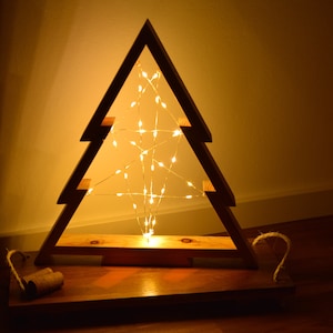 Könnte beinhalten: Eine Silhouette eines Weihnachtsbaums aus Holz mit einer Lichterkette aus warmweißen Lichtern im Inneren. Der Baum steht auf einem Holzsockel.