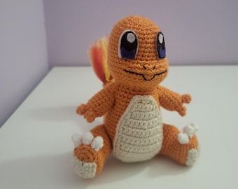 Puede incluir: Un peluche de Charmander naranja hecho a mano con ganchillo, con vientre color crema y garras blancas. El peluche tiene ojos negros grandes con detalles azules y una pequeña sonrisa. La cola es amarilla y roja. El juguete está sobre una superficie blanca.