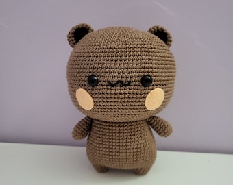 Può includere: Un orsetto all'uncinetto marrone fatto a mano con occhi neri e piccole orecchie. L'orso ha guance color beige e un piccolo sorriso. L'orso è in piedi su una superficie bianca contro uno sfondo viola. Questo peluche è perfetto per la cameretta di un bambino o come regalo.
