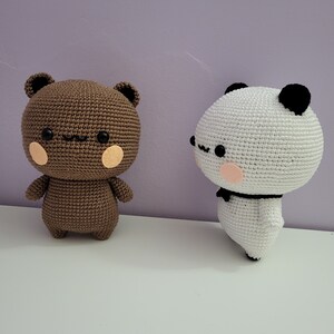 Puede incluir: Dos figuras de oso de ganchillo. Una es marr&oacute;n con ojos y orejas negras y mejillas de color rosa claro. La otra es blanca con orejas y ojos negros, un lazo negro y mejillas de color rosa claro. Ambas est&aacute;n sobre una superficie blanca.