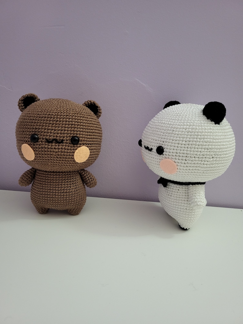 Puede incluir: Dos figuras de oso de ganchillo. Una es marr&oacute;n con ojos y orejas negras y mejillas de color rosa claro. La otra es blanca con orejas y ojos negros, un lazo negro y mejillas de color rosa claro. Ambas est&aacute;n sobre una superficie blanca.