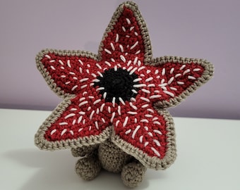 Puede incluir: Un peluche Demogorgon hecho a mano con ganchillo. La criatura presenta una cabeza roja en forma de estrella con detalles cosidos en blanco y un centro negro. La cabeza está delineada con un borde marrón. El cuerpo y los pies pequeños son de color marrón a juego.