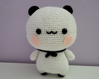 May include: A handmade white crochet panda bear doll with black eyes, ears, and feet. The panda has pink cheeks and a black bow tie. The doll is standing on a white surface against a purple background.