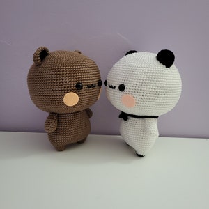 Puede incluir: Dos figuras amigurumi de ganchillo: un oso marr&oacute;n y un panda blanco. El oso tiene una cabeza redonda, orejas peque&ntilde;as y una mejilla color melocot&oacute;n. El panda tiene orejas negras, un lazo negro y una mejilla color melocot&oacute;n. Ambas figuras est&aacute;n juntas.