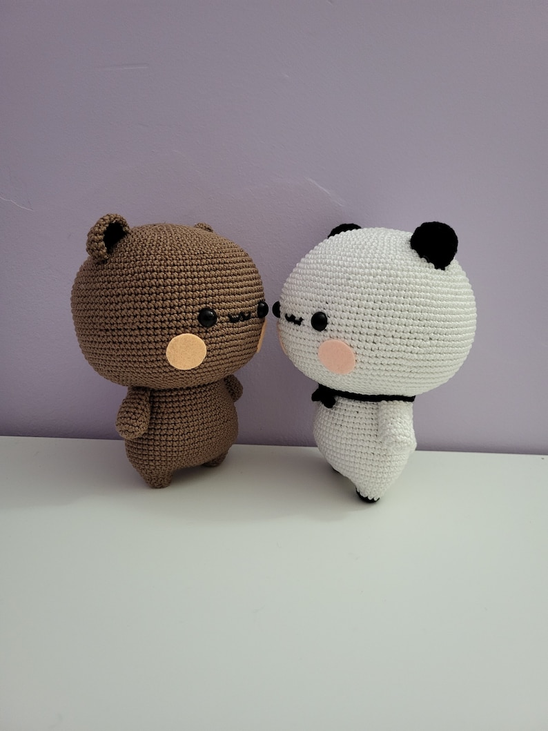 Puede incluir: Dos figuras amigurumi de ganchillo: un oso marr&oacute;n y un panda blanco. El oso tiene una cabeza redonda, orejas peque&ntilde;as y una mejilla color melocot&oacute;n. El panda tiene orejas negras, un lazo negro y una mejilla color melocot&oacute;n. Ambas figuras est&aacute;n juntas.