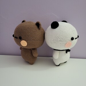 Puede incluir: Dos figuras amigurumi de ganchillo: un oso marr&oacute;n y un panda blanco. Ambos tienen gafas negras, mejillas rosadas y llevan una pajarita negra. El oso tiene orejas peque&ntilde;as y el panda tiene manchas negras en la cabeza.