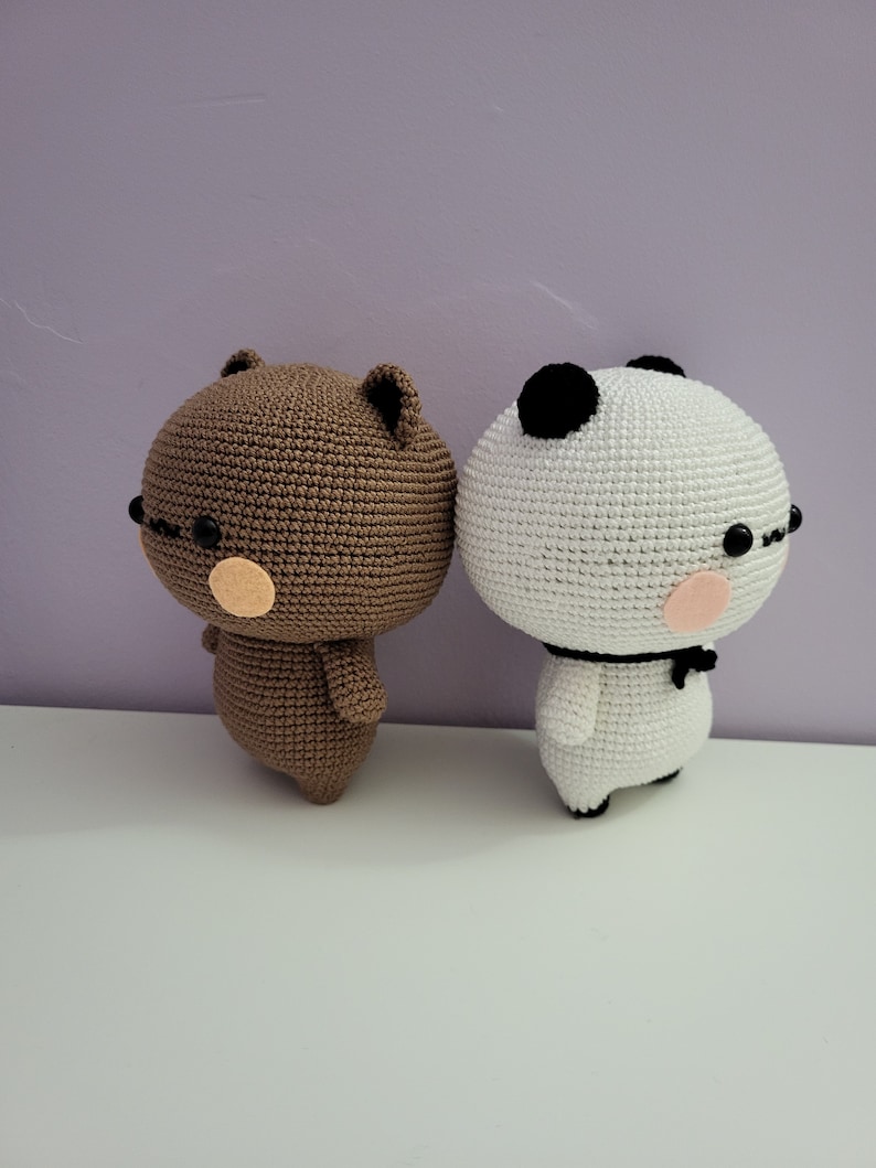 Puede incluir: Dos figuras amigurumi de ganchillo: un oso marr&oacute;n y un panda blanco. Ambos tienen gafas negras, mejillas rosadas y llevan una pajarita negra. El oso tiene orejas peque&ntilde;as y el panda tiene manchas negras en la cabeza.