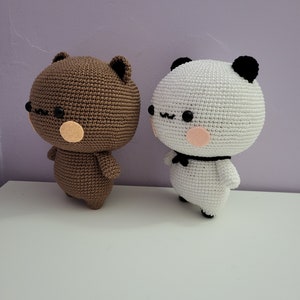 Puede incluir: Dos figuras de oso hechas a mano con ganchillo. Una es marr&oacute;n con detalles beige, la otra es blanca con orejas negras y una pajarita. Ambas tienen ojos negros y mejillas rosas. Est&aacute;n sobre una superficie blanca contra un fondo morado.