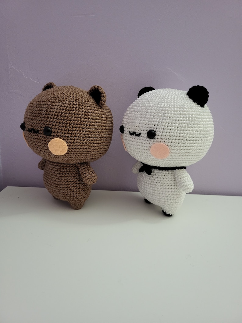 Puede incluir: Dos figuras de oso hechas a mano con ganchillo. Una es marr&oacute;n con detalles beige, la otra es blanca con orejas negras y una pajarita. Ambas tienen ojos negros y mejillas rosas. Est&aacute;n sobre una superficie blanca contra un fondo morado.