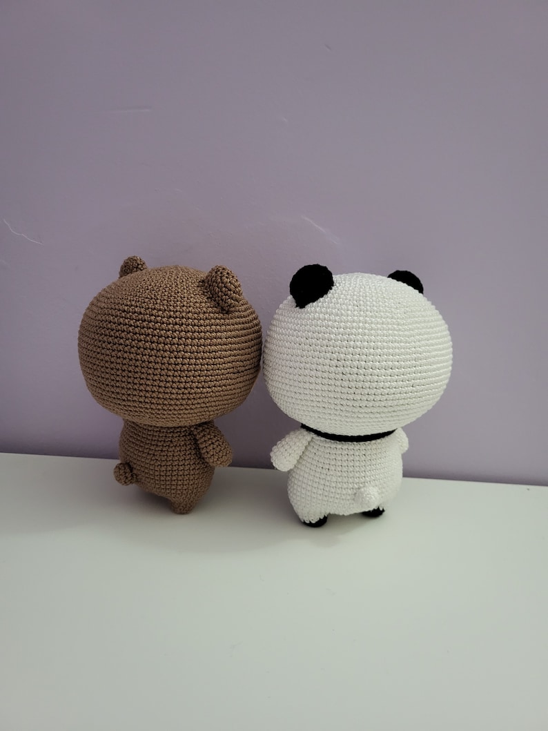 Puede incluir: Dos figuras de ganchillo hechas a mano, un oso y un panda. El oso es marr&oacute;n, y el panda es blanco con detalles negros en las orejas y un collar negro. Ambas figuras tienen orejas peque&ntilde;as y redondeadas y una peque&ntilde;a cola. Est&aacute;n sobre una superficie blanca contra un fondo morado.