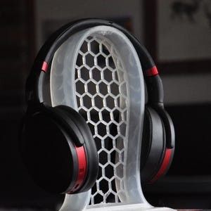 May include: Black over-ear headphones with red accents on the earcups, sitting on a white plastic stand with a honeycomb pattern.