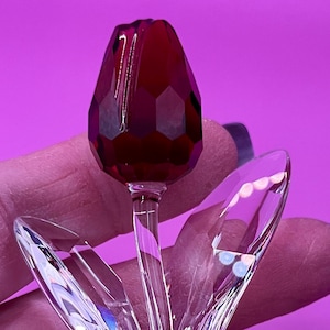 May include: A clear crystal tulip sculpture with a faceted, deep red flower head. The stem and leaves are transparent, reflecting light. The sculpture is held against a pink background, highlighting the intricate details of the glasswork.