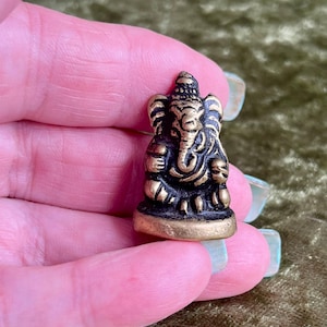 May include: A small, metallic, gold-toned figurine of the deity Ganesha in a seated position. The figure features a prominent elephant head, large ears, and a trunk curled to the left, with dark, recessed detailing that highlights the sculpted form.