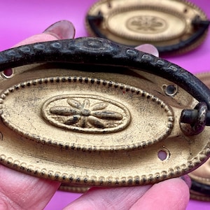 May include: Antique brass drawer pulls with a decorative floral design in the centre. The pulls have a gold finish with a dark, textured handle. The oval-shaped pulls are likely for furniture restoration.
