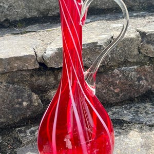 May include: A striking red glass jug with a clear glass handle. The jug has white, swirled stripes. It is displayed on a stone surface.