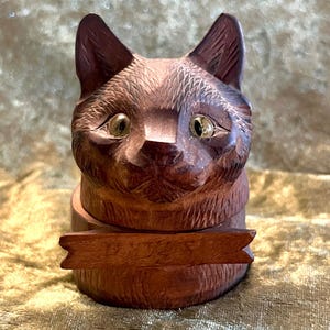 May include: A carved wooden cat figurine with a dark brown finish and light-coloured inset eyes. The cat head sits atop a cylindrical base, featuring a horizontal wooden banner across the front with the carved name Daisy in cursive script.