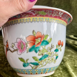 Puede incluir: Un cuenco de porcelana blanca con un diseño pintado a mano de un pequeño pájaro naranja posado en una rama con flores rosas y naranjas. El borde tiene una cenefa decorativa roja y amarilla, mientras que la base presenta un patrón geométrico.